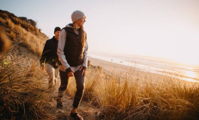 A senior couple explores a beach in Oregon state, enjoying the beauty of sunset on the Pacific Northwest coast.  They hike up a sand dune, the ocean visible in the background.